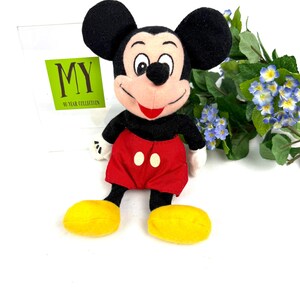 Vintage Mickey Mouse plush toy sitting upright with red shorts, yellow shoes, and flowers in the background.