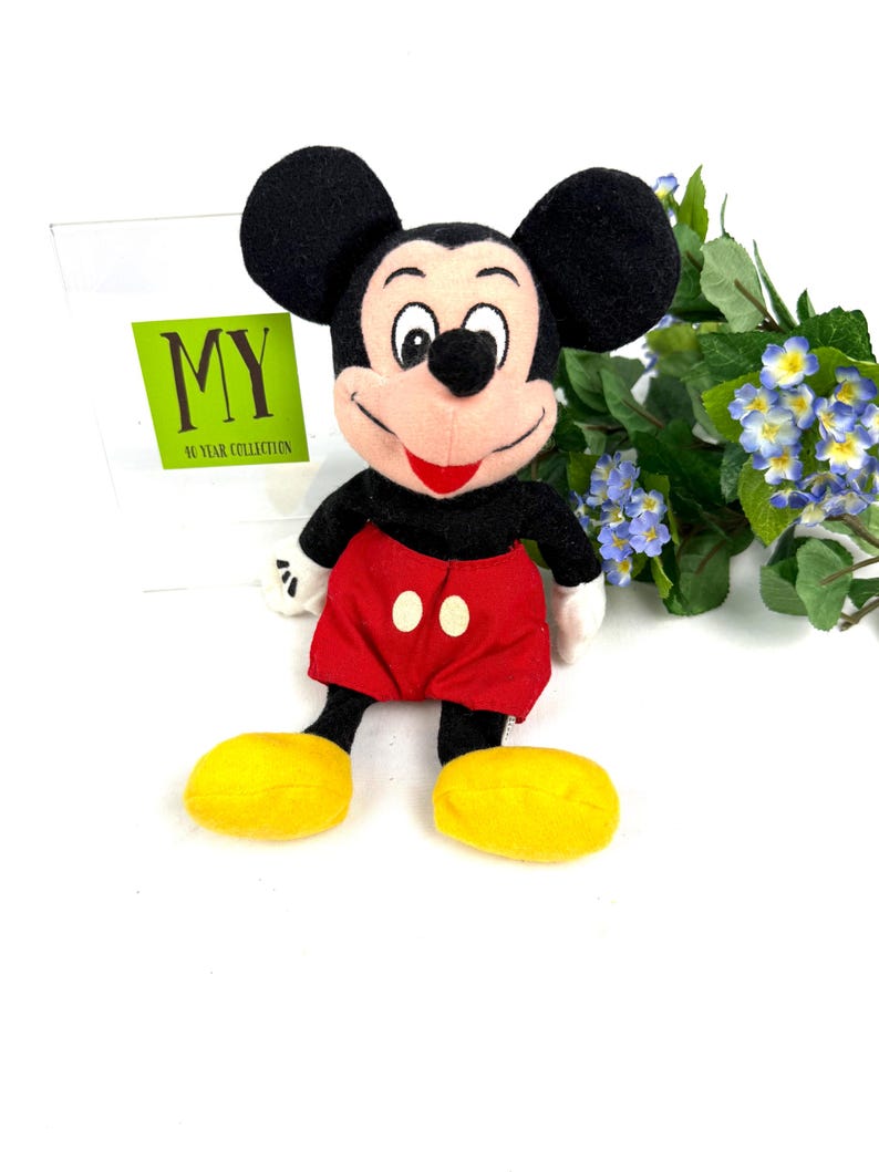 Vintage Mickey Mouse plush toy sitting upright with red shorts, yellow shoes, and flowers in the background.