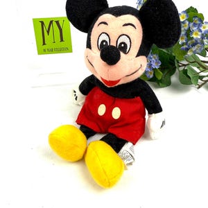 May include: A plush Mickey Mouse doll, featuring black ears, a black face, and a red mouth. The doll wears red shorts with two white buttons and yellow shoes. The text "MY 40 YEAR COLLECTION" is visible in the background.