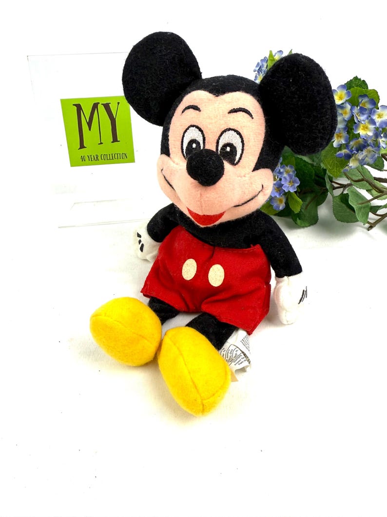 May include: A plush Mickey Mouse doll, featuring black ears, a black face, and a red mouth. The doll wears red shorts with two white buttons and yellow shoes. The text "MY 40 YEAR COLLECTION" is visible in the background.