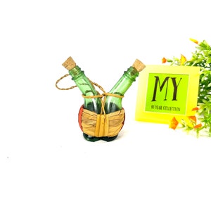 May include: Two small green glass bottles with cork tops are tied together with twine and placed in a woven basket. The basket is brown with red accents. The bottles are sitting on a white surface. The image also includes a yellow sign with the text "MY 40 YEAR COLLECTION".