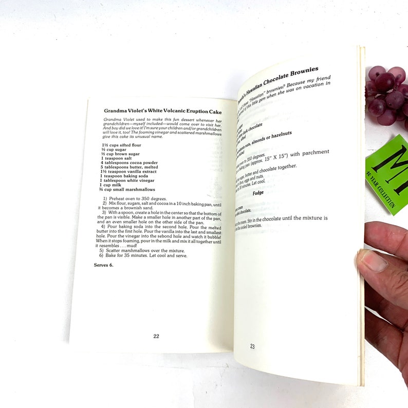 May include: A close-up of two open recipe books with a white background. The left page features a recipe for Grandma Violet's White Volcanic Eruption Cake, with instructions and ingredients listed. The right page features a recipe for Hawaiian Chocolate Brownies, with instructions and ingredients listed.