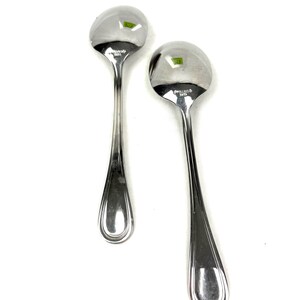 May include: Two shiny, silver spoons with rounded bowls and long, slender handles. The spoons are made of a reflective metal, possibly stainless steel, and are set against a white background. The spoons are likely for serving or eating.