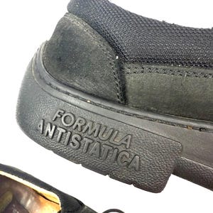 May include: Close-up of a black shoe with the words "FORMULA ANTISTATICA" embossed on the sole. The shoe has a textured rubber sole and a suede upper. The shoe's heel is made of a black mesh material.