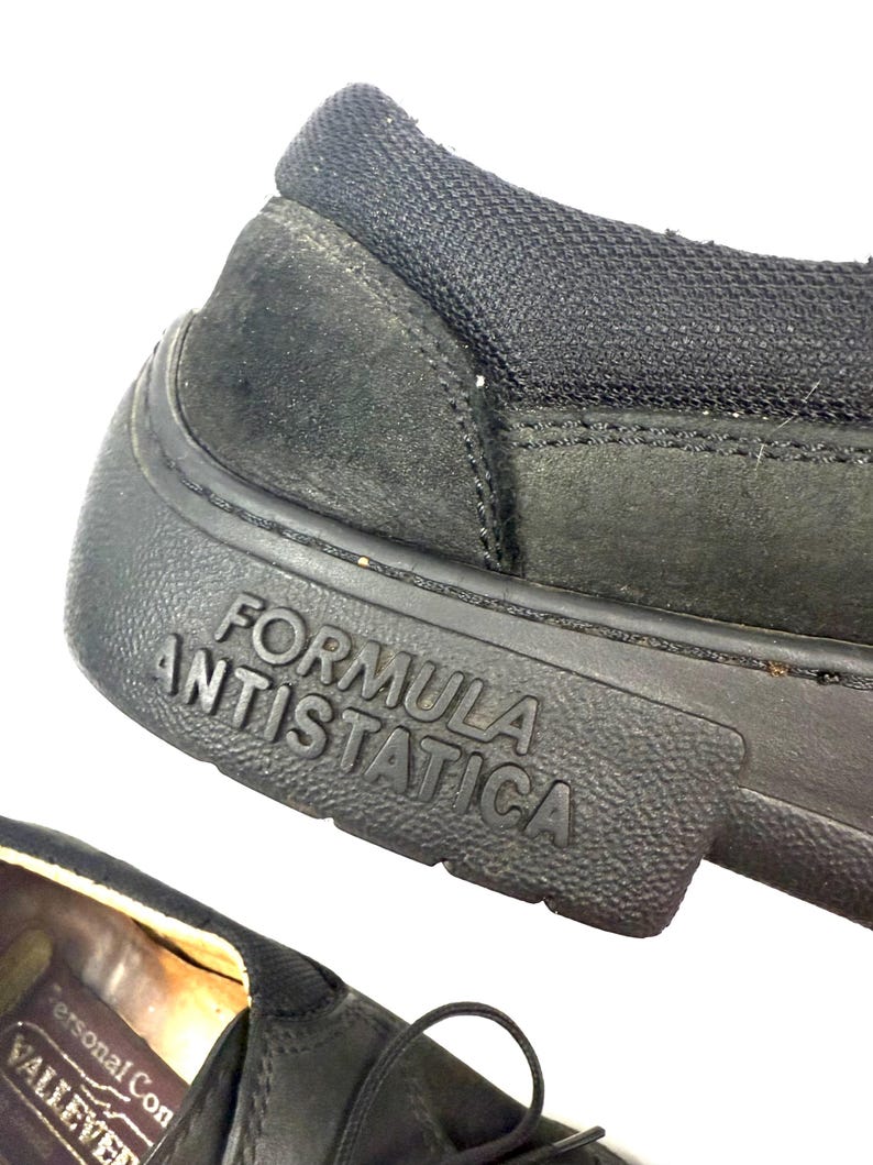 May include: Close-up of a black shoe with the words "FORMULA ANTISTATICA" embossed on the sole. The shoe has a textured rubber sole and a suede upper. The shoe's heel is made of a black mesh material.