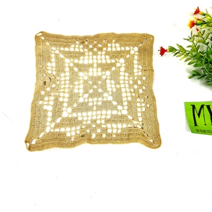 Op de afbeelding: Een vierkant, beige gehaakt tafellaken met een delicaat, ingewikkeld patroon. Het tafellaken is gemaakt van een fijn, lichtgewicht garen en heeft een zachte, luchtige textuur.