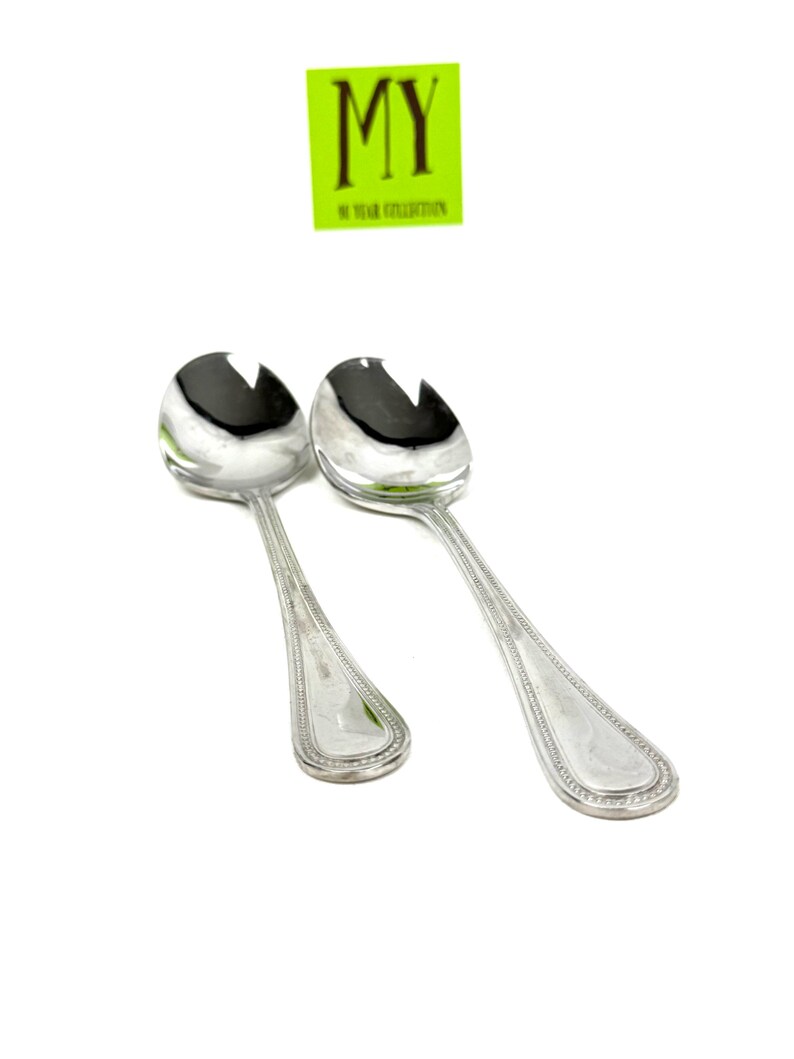 May include: Two silver serving spoons with slotted bowls and decorative handles. The spoons are angled towards the viewer against a white background. A green sticker with the text "MY 50 YEAR COLLECTION" is in the upper portion of the image.