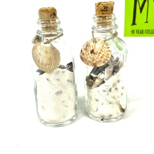 May include: Two small glass bottles with cork stoppers filled with white sand and seashells. Each bottle has a seashell tied to the neck with twine.