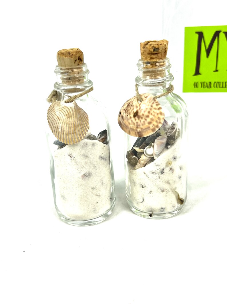 May include: Two small glass bottles with cork stoppers filled with white sand and seashells. Each bottle has a seashell tied to the neck with twine.