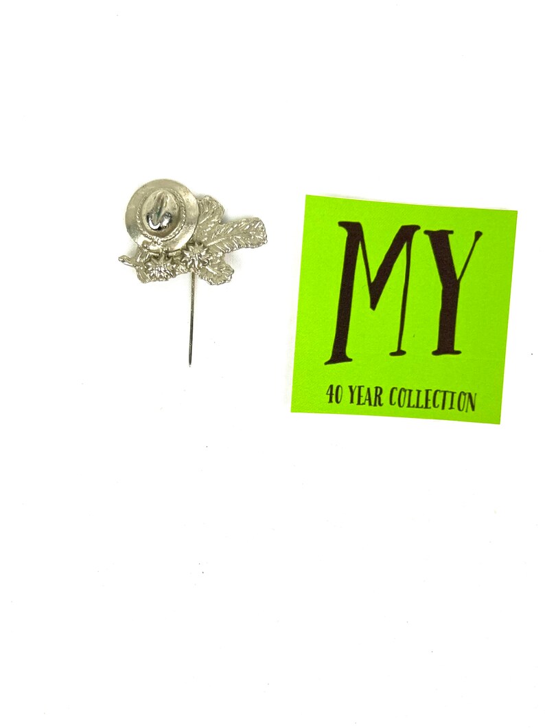 May include: Silver pin with a hat and leaves design. The pin is attached to a green card with the text "MY 40 YEAR COLLECTION".