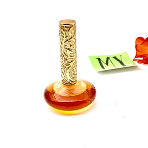 May include: A vintage amber glass perfume bottle with a gold, ornate, floral-patterned stopper. The bottle is sitting on a white surface.