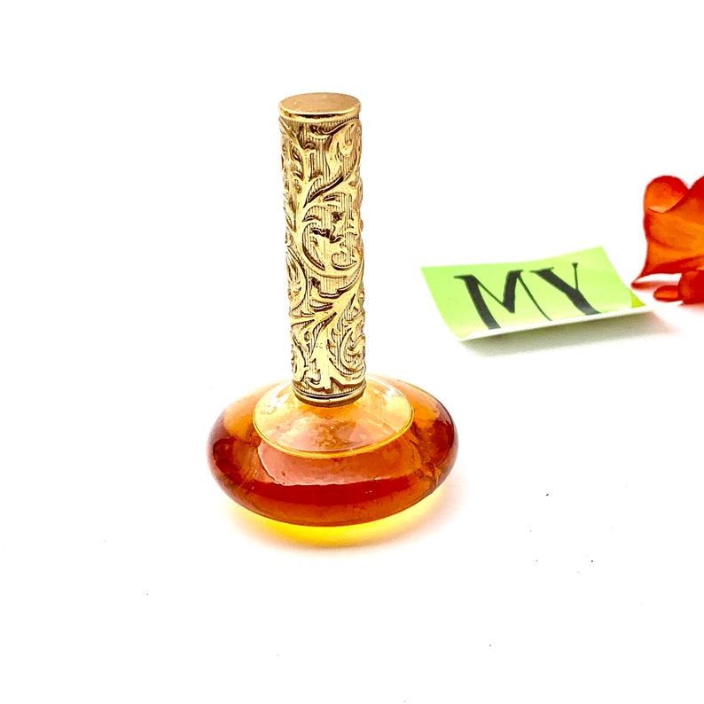 May include: A vintage amber glass perfume bottle with a gold, ornate, floral-patterned stopper. The bottle is sitting on a white surface.