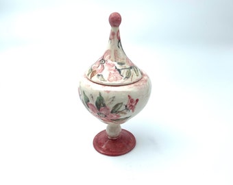 May include: A white ceramic jar with a pink floral design and a red lid. The jar has a round body and a pedestal base.
