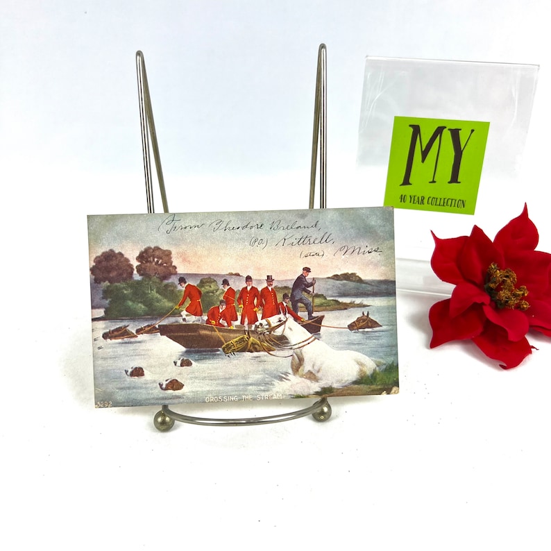 K&ouml;nnte beinhalten: Vintage-Postkarte, die eine Gruppe von M&auml;nnern in roten M&auml;nteln zeigt, die einen Fluss in einem Boot &uuml;berqueren, mit dem Text "From Theodore Breland, (80) Kittrell, Miss" und "CROSSING THE STREAM" auf der Postkarte.
