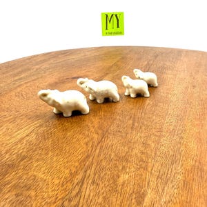 May include: Four small, off-white ceramic elephant figurines arranged on a wooden surface. The elephants are detailed with visible trunks, ears, and legs. The image includes a green and yellow sticker with the text "MY 96 YEAR COLLECTION".