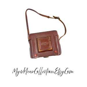 May include: A vintage brown leather camera case with a matching strap. The case has a square design with a central embossed detail and red button closures. The text "DUMAS PARIS" is visible on the case. The Etsy shop name "My4YearCollection.Etsy.Com" is at the bottom.