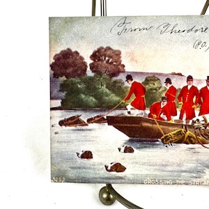 K&ouml;nnte beinhalten: Eine antike Postkarte, die eine Gruppe von M&auml;nnern in roten M&auml;nteln zeigt, die einen Fluss in einem Boot &uuml;berqueren. Die M&auml;nner ziehen an einem Seil, um das Boot zu lenken. Die Postkarte tr&auml;gt den Titel "Crossing the Stream" und die handschriftliche Unterschrift "Groom Theodore (P.O.)" in der oberen rechten Ecke.