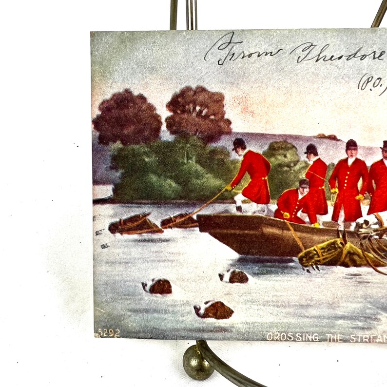 K&ouml;nnte beinhalten: Eine antike Postkarte, die eine Gruppe von M&auml;nnern in roten M&auml;nteln zeigt, die einen Fluss in einem Boot &uuml;berqueren. Die M&auml;nner ziehen an einem Seil, um das Boot zu lenken. Die Postkarte tr&auml;gt den Titel "Crossing the Stream" und die handschriftliche Unterschrift "Groom Theodore (P.O.)" in der oberen rechten Ecke.