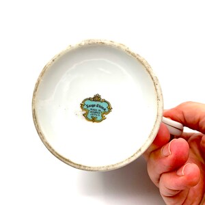 以下が含まれることがあります： 白磁のカップの底面。青と金のラベルには「Sango China Made in Occupied Japan」と記載されています。