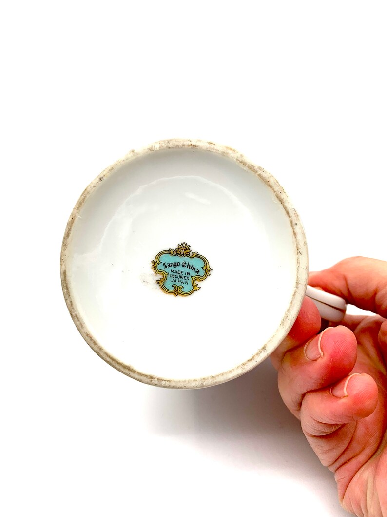 以下が含まれることがあります： 白磁のカップの底面。青と金のラベルには「Sango China Made in Occupied Japan」と記載されています。