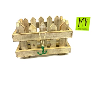 Könnte beinhalten: Ein kleiner, rustikaler Pflanzkasten im Zaun-Stil aus hellem Holz mit verwittertem Aussehen. Ein grüner, dekorativer Anker hängt von der Vorderseite. Der Text "MY 10 YEAR COLLECTION" befindet sich auf einem grünen Aufkleber.
