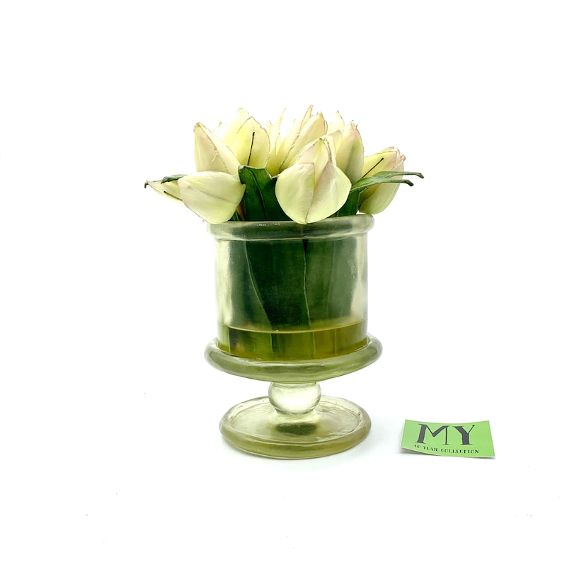 May include: A white glass vase with a pedestal base holds a bouquet of white tulips with green leaves. The vase has a clear glass body and a textured, frosted base. The vase is sitting on a white surface.