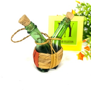 May include: Two small green glass bottles with cork stoppers are tied together with twine and placed in a woven basket with a red stripe. The bottles are in a decorative display.