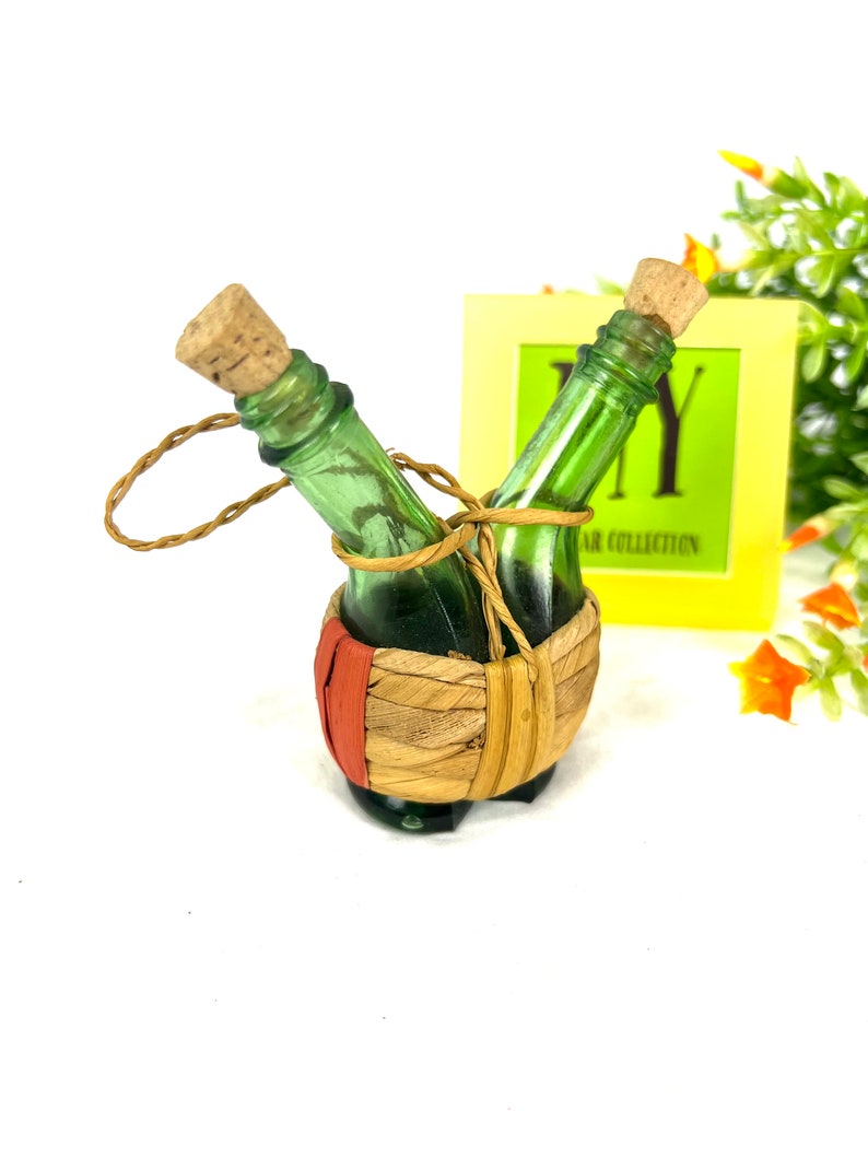 May include: Two small green glass bottles with cork stoppers are tied together with twine and placed in a woven basket with a red stripe. The bottles are in a decorative display.