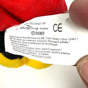 May include: A close-up of a Disney product tag. The tag is white with black text, including the Disney logo and the text "MADE EXCLUSIVELY FOR". The tag also includes the CE mark and manufacturing information. The product is red, yellow, and black.