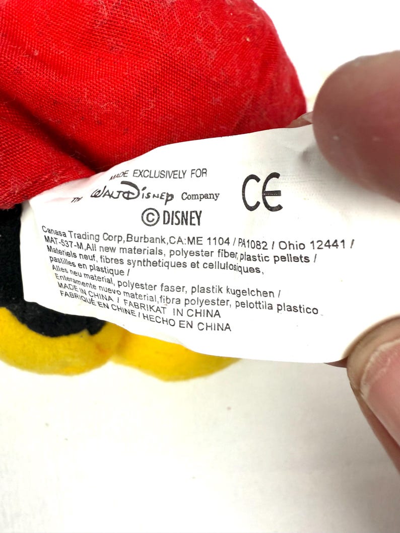 May include: A close-up of a Disney product tag. The tag is white with black text, including the Disney logo and the text "MADE EXCLUSIVELY FOR". The tag also includes the CE mark and manufacturing information. The product is red, yellow, and black.