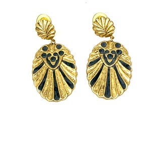 Könnte beinhalten: Ein Paar goldfarbene Ohrringe mit muschelartigem Design. Jeder Ohrring hat eine kleine, gewellte Oberseite und einen ovalen Anhänger mit schwarzen Emaille-Akzenten. Die Ohrringe haben eine Vintage-Ästhetik.