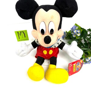 May include: A plush Mickey Mouse toy with black ears, a tan face, and a red outfit with yellow buttons. The toy has white gloves and large yellow feet. A tag is attached to the toy.