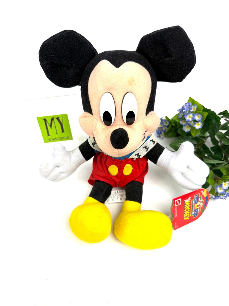 May include: A plush Mickey Mouse toy with black ears, a tan face, and a red outfit with yellow buttons. The toy has white gloves and large yellow feet. A tag is attached to the toy.
