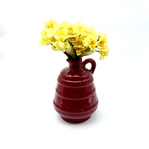 Könnte beinhalten: Eine rote Keramikkanne mit einem ausgestellten Ausguss und einem Henkel. Die Kanne ist mit horizontalen Rillen verziert und mit einem Strauß gelber Blumen gefüllt.