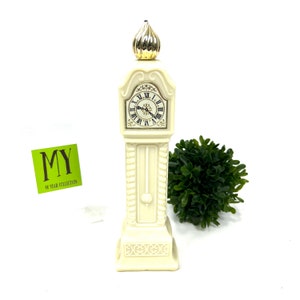 May include: A white ceramic clock shaped like a grandfather clock with gold accents and a black clock face. The clock is on a white surface.