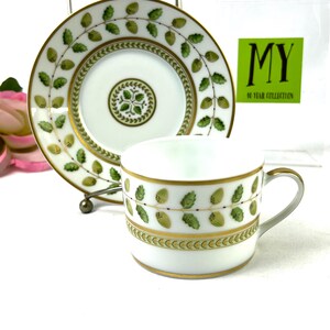 May include: A white porcelain teacup and saucer with a green and gold floral pattern. The saucer has a gold rim and the cup has a gold band around the top. The pattern features green leaves and gold berries.