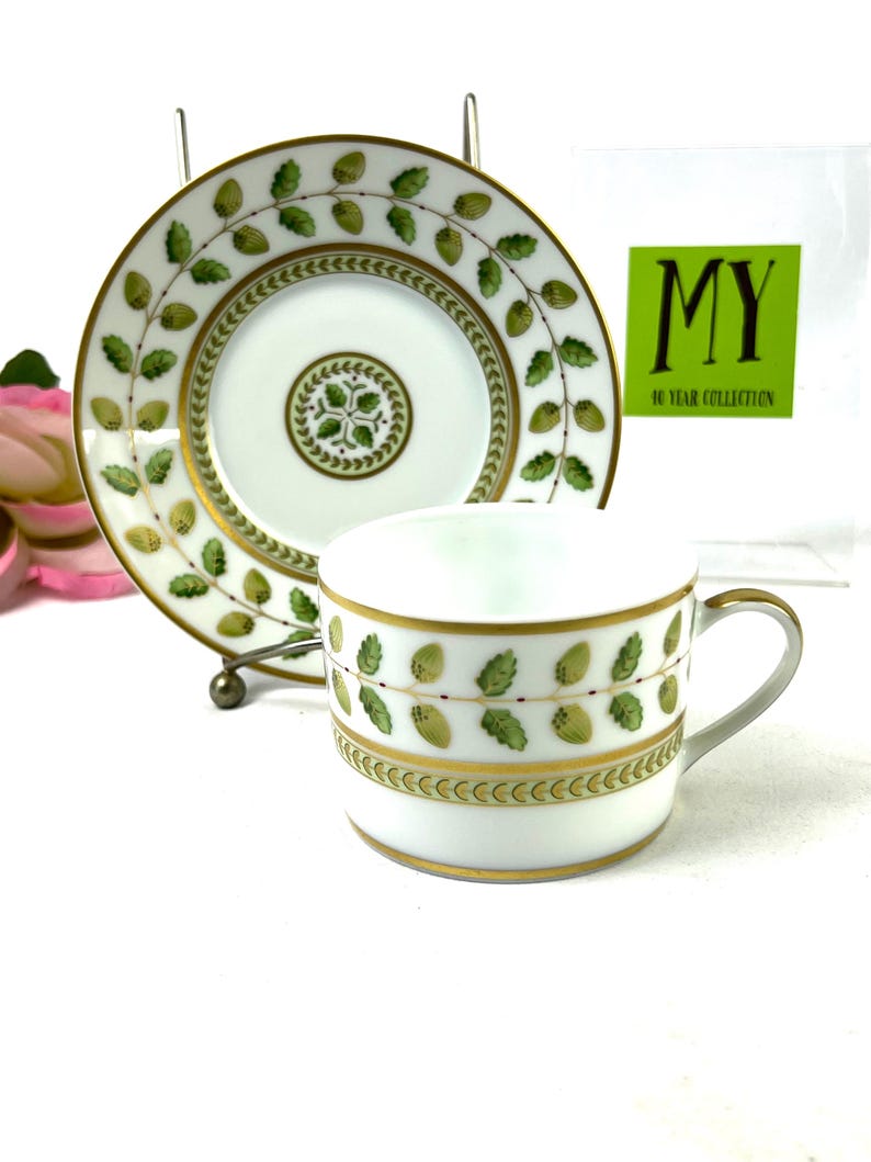 May include: A white porcelain teacup and saucer with a green and gold floral pattern. The saucer has a gold rim and the cup has a gold band around the top. The pattern features green leaves and gold berries.
