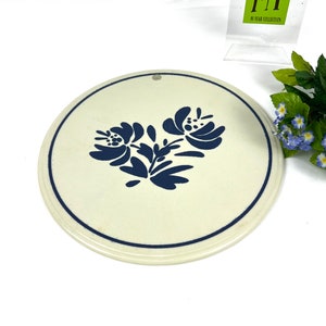 May include: A white ceramic trivet with a blue floral design. The trivet is round and has a hole in the centre for hanging.
