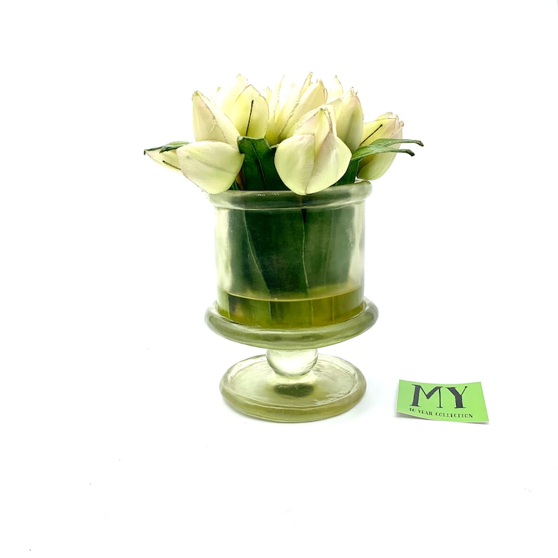 May include: A clear glass vase with a pedestal base holds a bouquet of white tulips. The vase has a textured surface and the tulips have green leaves.