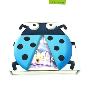 Puede incluir: Una caja azul en forma de mariquita con puntos negros y ojos blancos. La caja está abierta para revelar un interior blanco con un diseño floral y una pequeña mariquita roja.