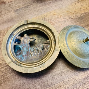 May include: A brass ashtray with a lid. The ashtray features a raised design of a religious scene with figures in the centre. The lid has a floral design.