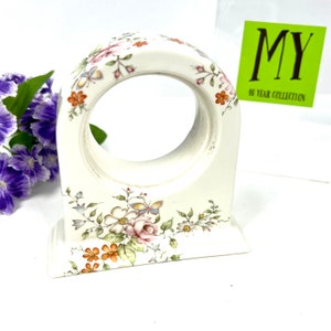 May include: White ceramic clock stand with a floral design featuring pink, orange, and green flowers and butterflies. The stand has a circular opening in the center.