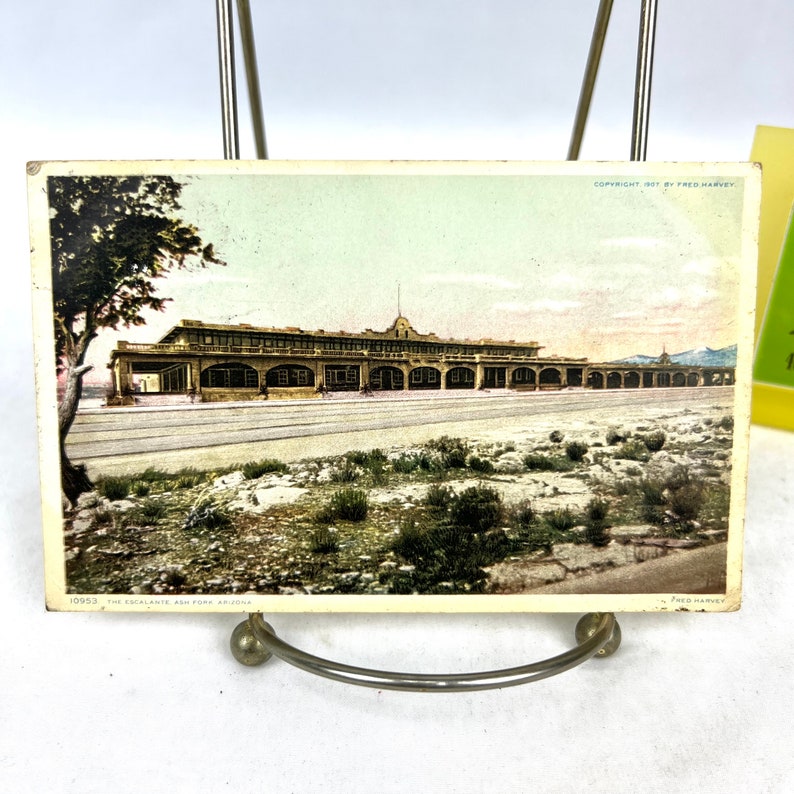 May include: A vintage postcard depicting the Escalante Ash Fork in Arizona, showing a long building with a covered walkway. The postcard has a faded colour palette, a desert landscape, and the text "COPYRIGHT 1907 BY FRED HARVEY."