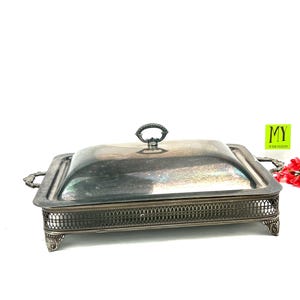 May include: Silver serving dish with a lid and a decorative base. The dish has a square shape and a raised rim. The lid has a knob on top.