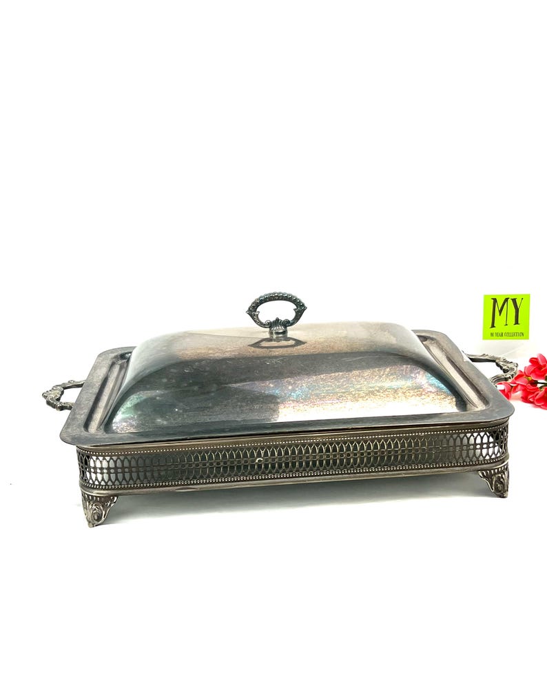 May include: Silver serving dish with a lid and a decorative base. The dish has a square shape and a raised rim. The lid has a knob on top.