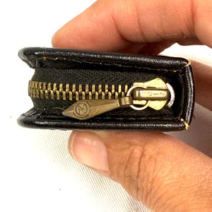 May include: A small, black leather coin purse with a brass zipper. The zipper is partially open, revealing the interior. The zipper pull has an "N" logo. The purse is held by a person's hand.