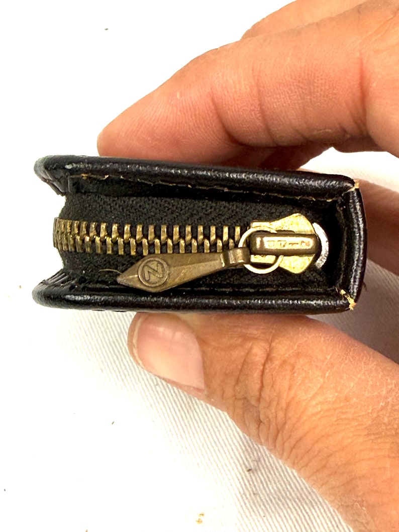 May include: A small, black leather coin purse with a brass zipper. The zipper is partially open, revealing the interior. The zipper pull has an "N" logo. The purse is held by a person's hand.