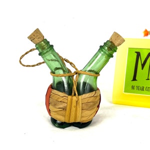 May include: Two small green glass bottles with cork stoppers are tied together with twine and placed in a woven basket with red and orange accents.