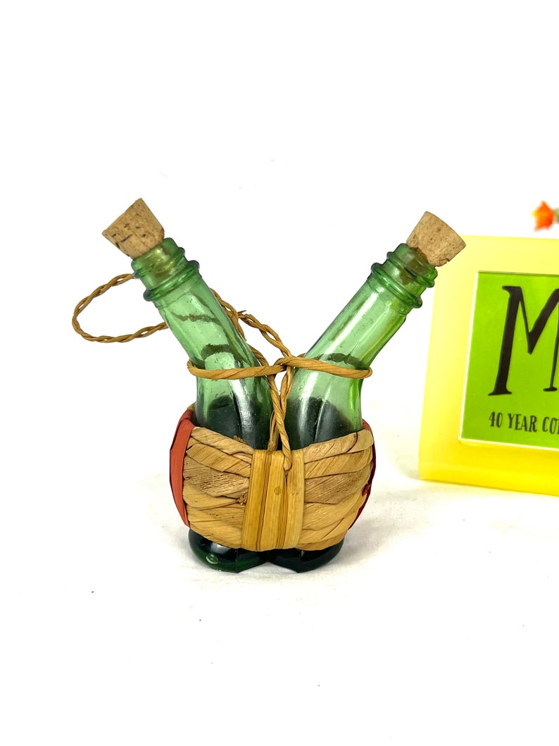 May include: Two small green glass bottles with cork stoppers are tied together with twine and placed in a woven basket with red and orange accents.