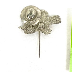 May include: Silver-toned pin featuring a stylized hat with a flower design. The pin is attached to a long, thin pin.